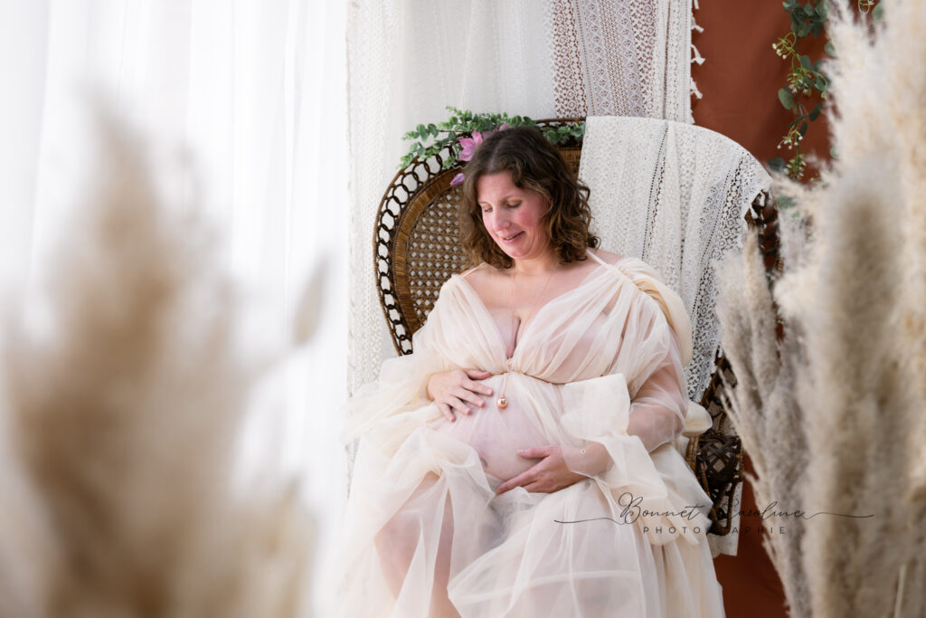 Séance photo grossesse en studio à Morez : future maman assise dans un fauteuil en rotin, robe en voile rose