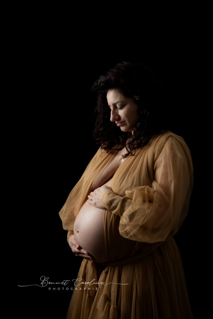 Séance photo grossesse en studio à Morez : future maman en robe brune sur fond noir, regard vers son ventre