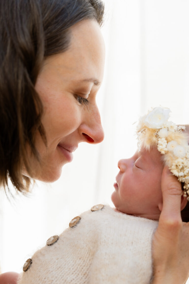 Séance photo naissance en studio à Morez : maman qui sourit à son bébé endormi coiffé d’une couronne de fleurs