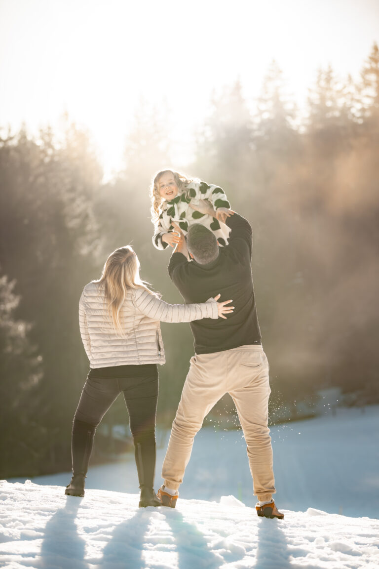 Séance photo famille dans le Haut-Jura : parents faisant tournoyer leur enfant dans la neige au soleil