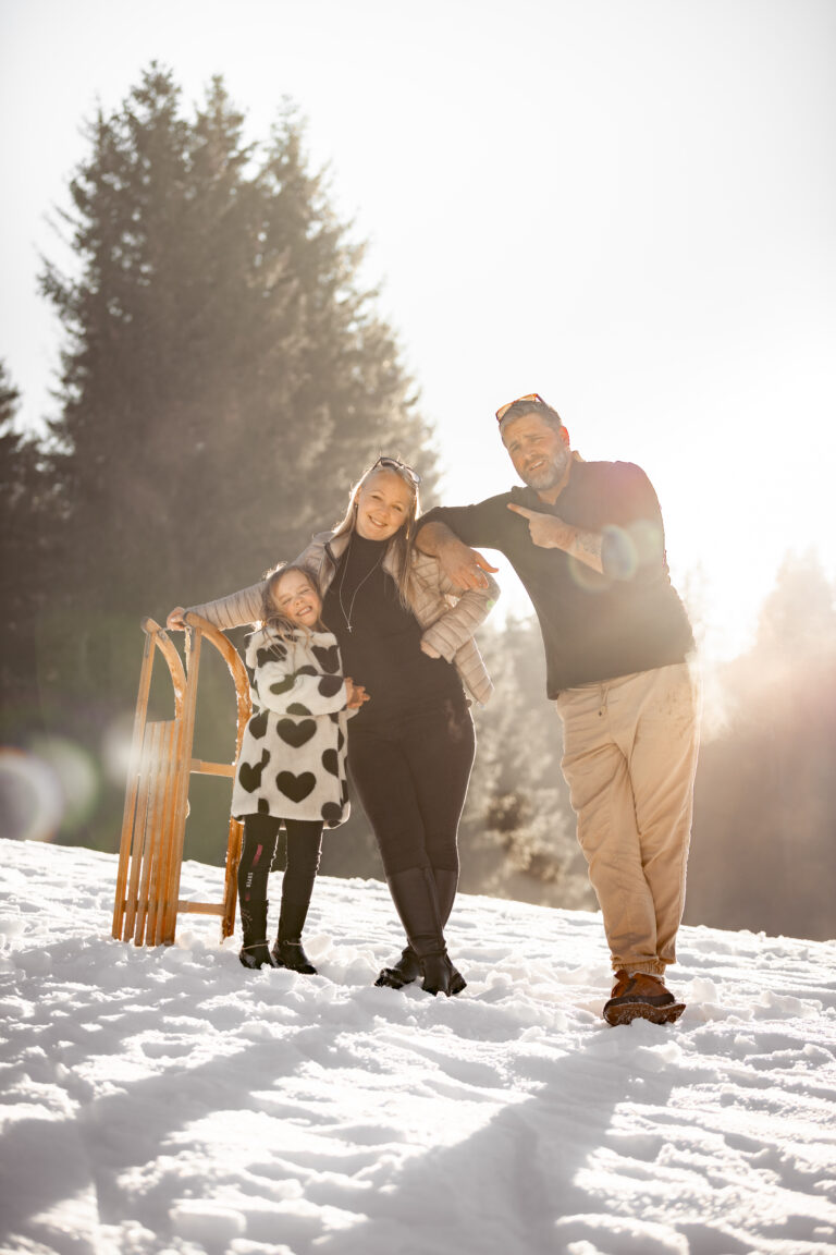 Séance photo famille dans le Haut-Jura : famille debout près d’une luge dans la neige au soleil