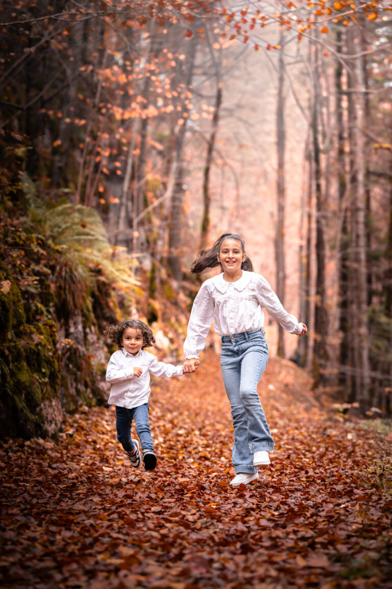 Séance photo famille dans le Haut-Jura : deux sœurs courant main dans la main sur un tapis de feuilles d’automne