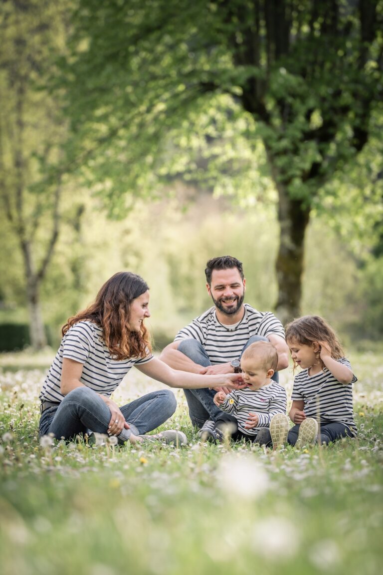Séance photo famille dans le Haut-Jura : parents et leurs enfants assis dans l’herbe sous les arbres