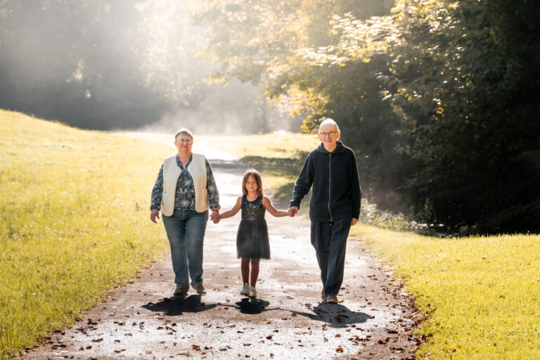 Séance photo famille dans le Haut-Jura : grands-parents et leur petite-fille se promenant sur un chemin à la lumière douce