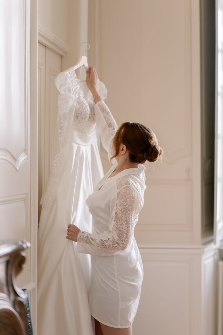 Photographe mariage Jura : mariée qui décroche sa robe près de la fenêtre