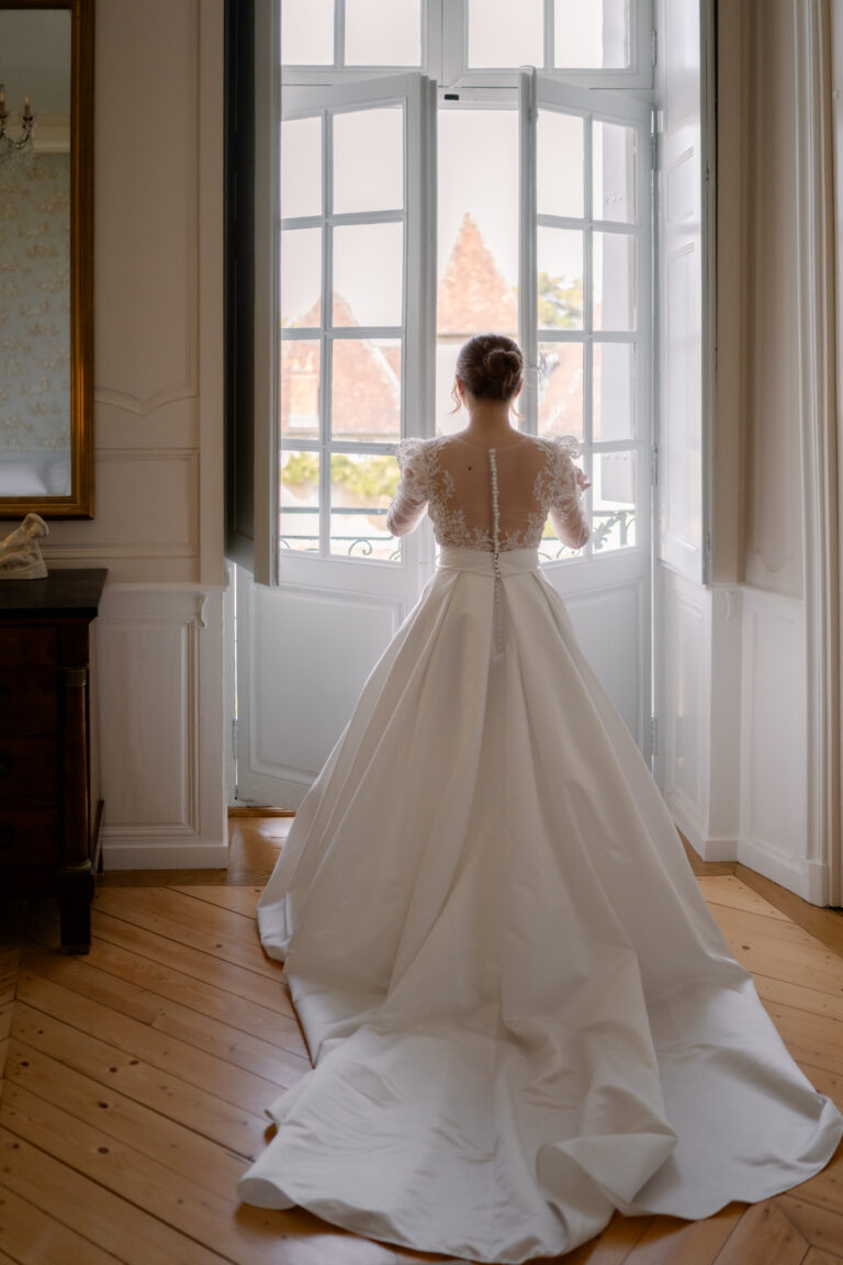 Photographe mariage Jura : mariée de dos devant la fenêtre, longue traîne