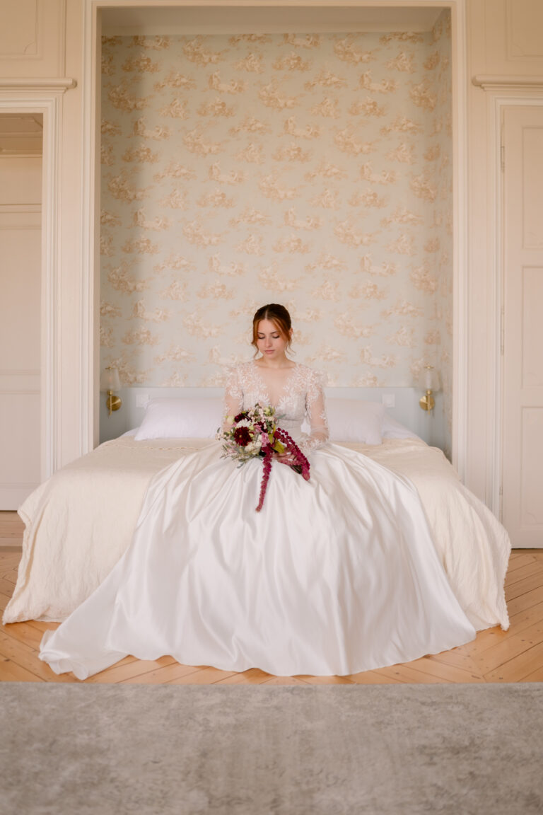 Photographe mariage Jura : mariée assise sur le lit, robe étalée et bouquet bordeaux