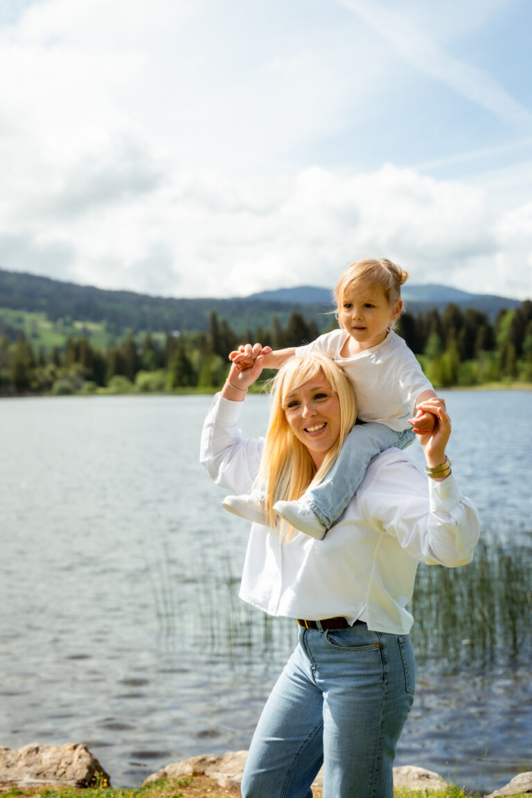 Séance photo famille dans le Haut-Jura : maman portant sa fille sur les épaules au bord d’un lac