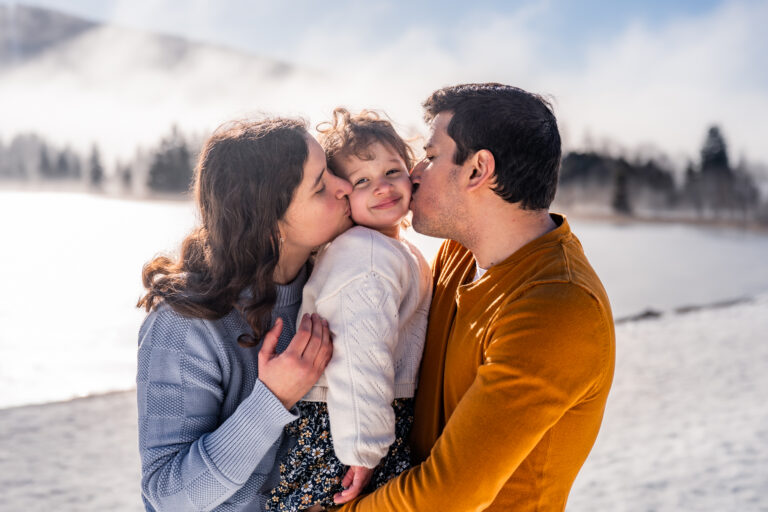 Séance photo famille dans le Haut-Jura : parents embrassant leur enfant au bord d’un lac enneigé