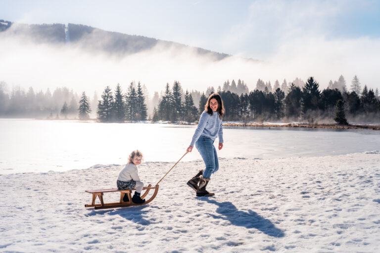 Séance photo famille dans le Haut-Jura : maman tirant sa fille en luge au bord d’un lac enneigé