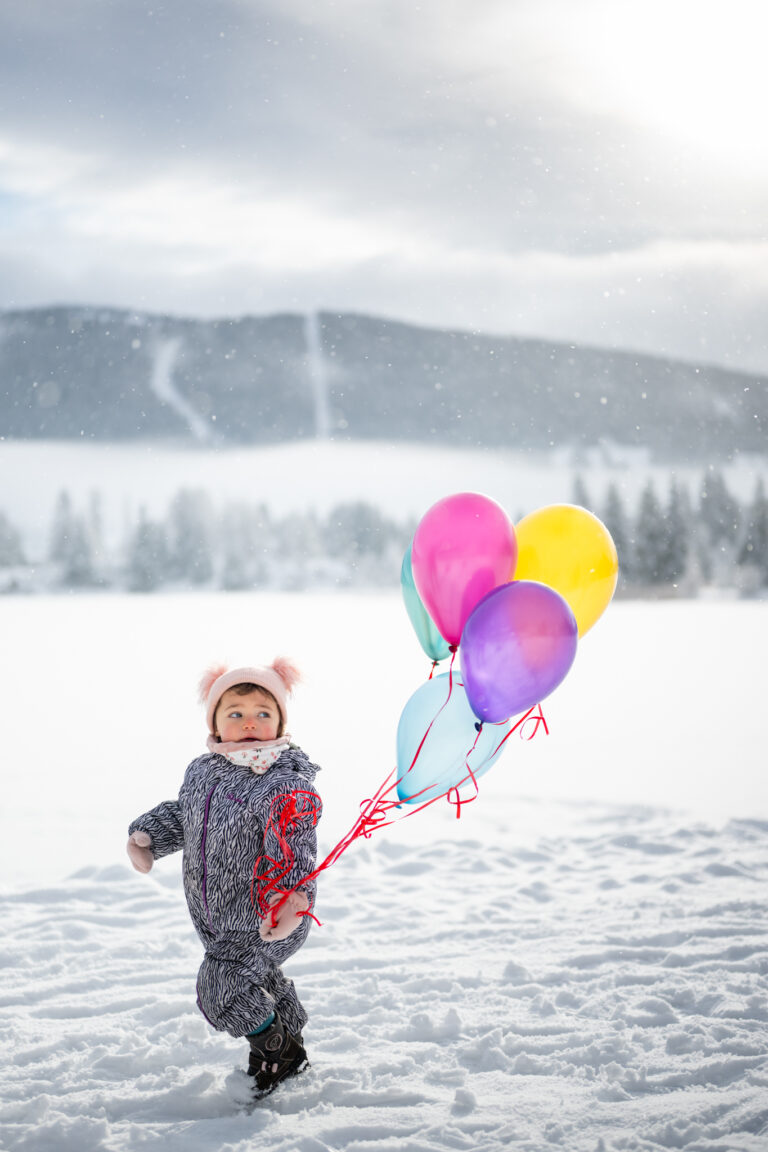 Séance photo famille dans le Haut-Jura : petite fille marchant dans la neige avec des ballons colorés