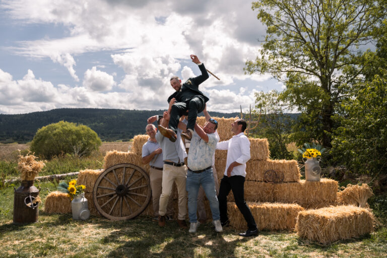 Photographe mariage Jura : amis qui font sauter le marié dans les airs près des bottes de paille