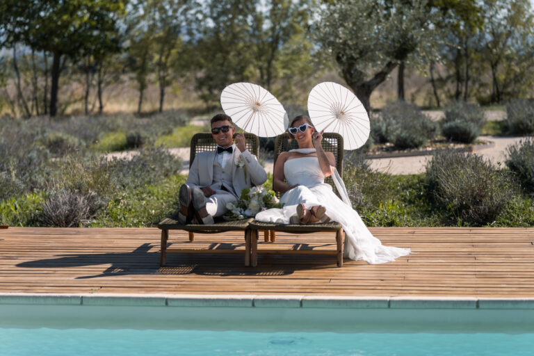 Photographe mariage Jura : mariés en lunettes de soleil sur des transats au bord de la piscine, ombrelles blanches, dans la Drôme