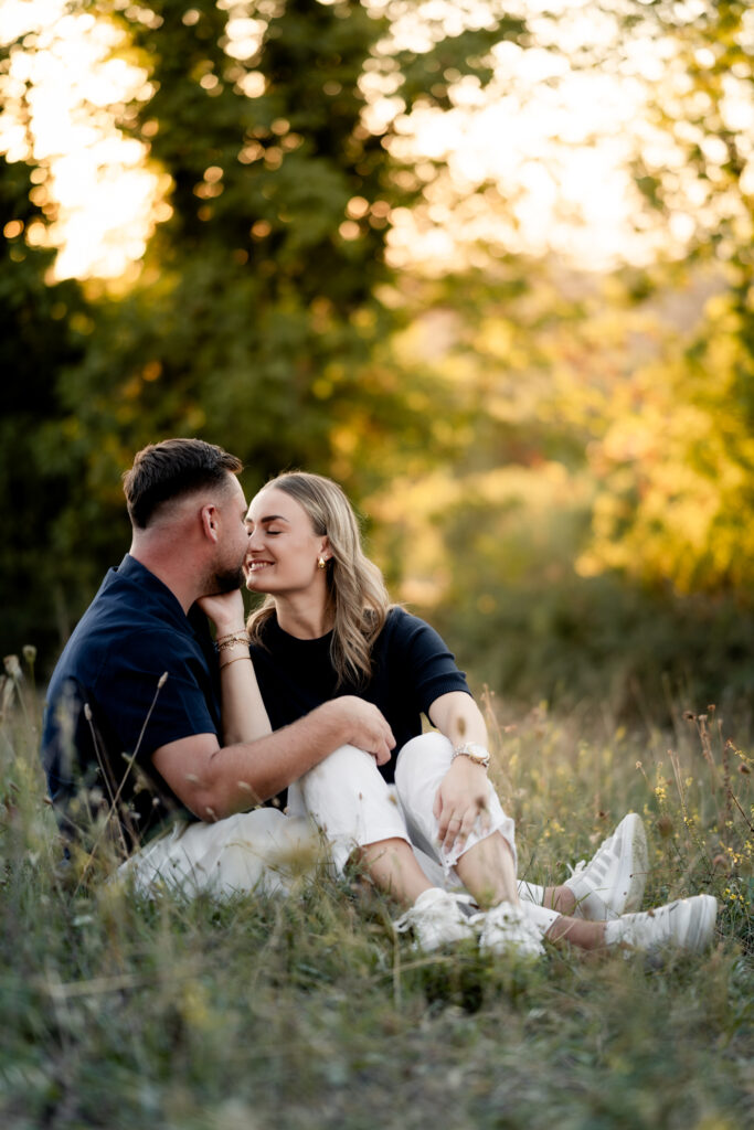 Couple sitting in the grass and kissing in soft evening light, natural engagement photography in the Jura, near Lausanne