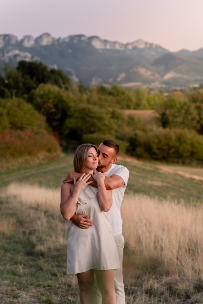 Couple embracing in a golden meadow with mountain cliffs in the background, engagement photo session in the Jura, near Switzerland