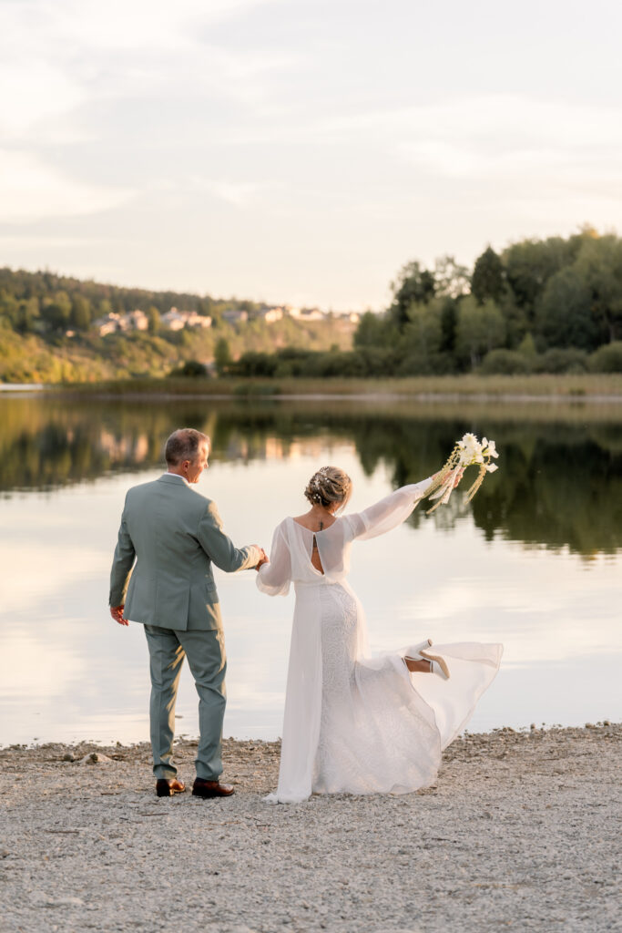 Bride raising her bouquet and twirling her dress next to her groom on the shore of Lac de l'Abbaye at sunset, Jura, France