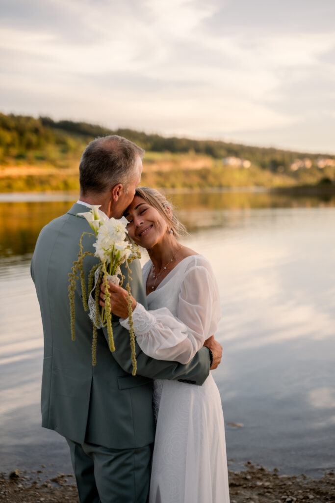 Photographe mariage Jura : mariée avec son bouquet blanc, tête contre l’épaule de son marié au bord du lac de l’Abbaye
