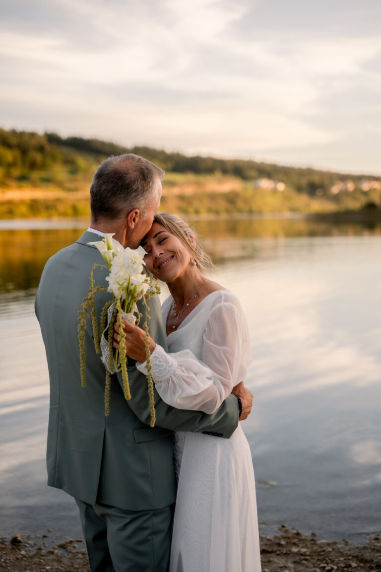 Photographe mariage Jura : mariée avec son bouquet blanc, tête contre l’épaule de son marié au bord du lac de l’Abbaye