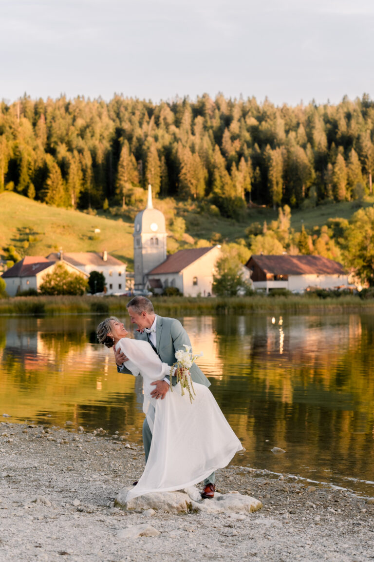 Photographe mariage Jura : mariée qui danse au bord du lac de l’Abbaye, l’église en arrière-plan