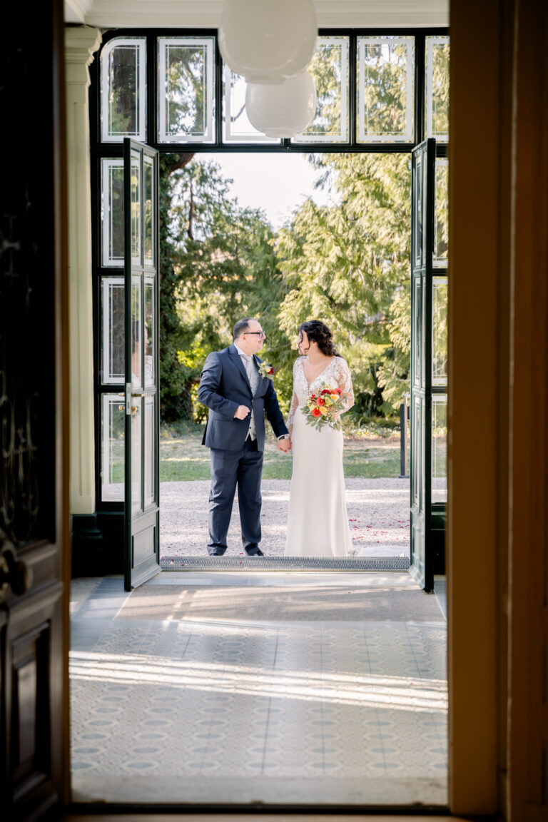 Photographe mariage Jura : mariés vus à travers la grande porte vitrée du château
